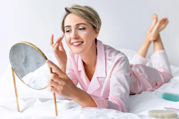 Roześmiana kobieta leży na łóżku i przed małym lusterkiem aplikuje krem pod oczy 